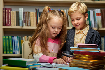 school kids reading book together while sitting at table in library, boy and girl among lots of books, preparing fro school