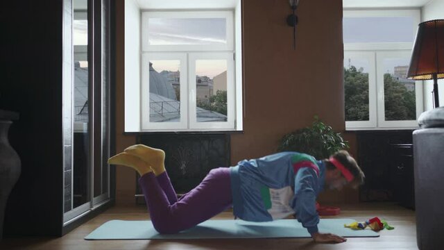 Funny whimsical guy lying on a fitness mat, doing push-ups from the floor, dressed in 80s style in a bright sports suit, the guy doing fitness at home on self-isolation.