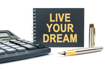 On a white background, there is a calculator, a pen and a black notebook with the inscription - LIVE YOUR DREAM