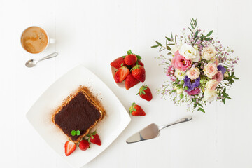 Tiramisu, homemade Italian sweet no bake cheesecake dessert, on white plate embellished with fresh mint and a few real strawberries, cup of coffee, bowl with strawberries and flowers on white table.
