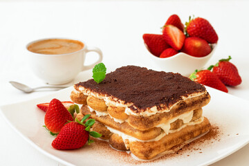 Tiramisu, homemade sweet Italian no bake dessert, cut of cake on small plate embellished with fresh mint and a few real strawberries, cup of coffee and bowl with strawberries on white table. Closeup.