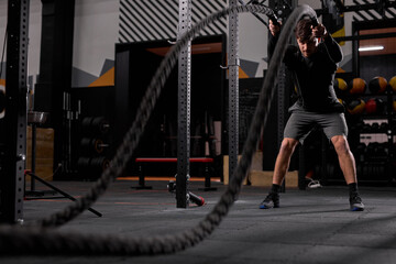 Fit athlete doing battle rope exercise at gym, having intense training alone. Concentrated...
