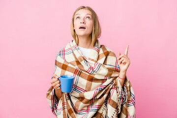 Young russian woman wrapped in a blanket drinking coffee pointing upside with opened mouth.