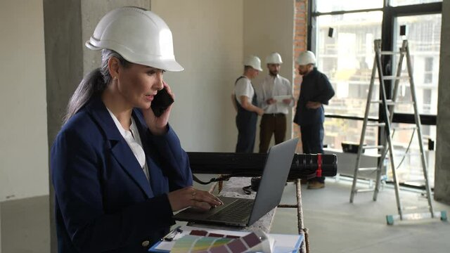 Female architect in helmet talking on smartphone while working on laptop pc in apartment under renovation. Designer busy on phone at work, male architect giving task to builders on background