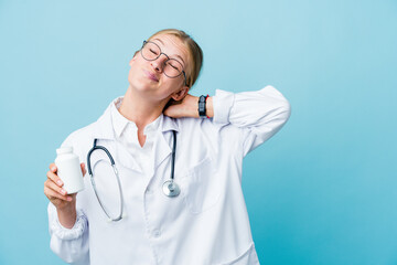 Young russian doctor woman holding pills bottle on blue touching back of head, thinking and making a choice.