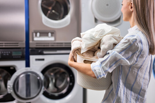 woman doing laundry at laundry room with washing machine, holding basket with linen and towels in hands. modern technologies, cleaning and washing concept