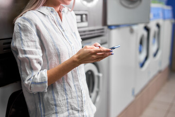 Enjoying of easy laundry process. cropped photo of young lady standing near washing machine and looking at smartphone app, waiting end of washing