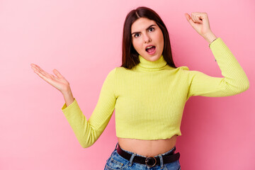 Young caucasian woman isolated on pink background holds copy space on a palm, keep hand over cheek. Amazed and delighted.