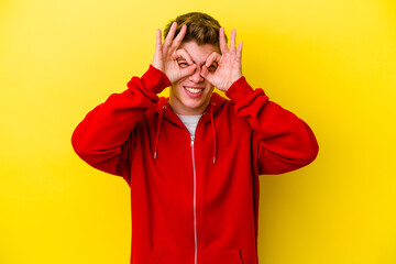 Young caucasian man isolated on yellow background showing okay sign over eyes