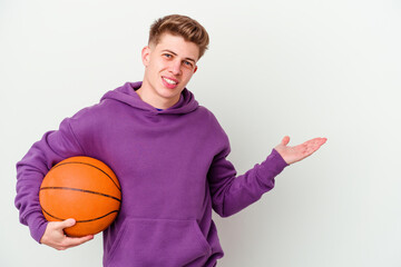 Young caucasian man playing basketball isolated background showing a copy space on a palm and holding another hand on waist.