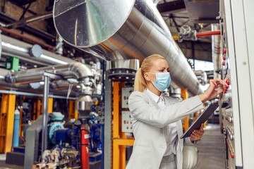 Dedicated female blond supervisor with face mask standing in heating plant next to dashboard, adjusting settings and holding tablet during corona virus pandemic.