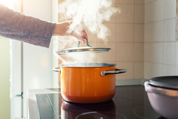 Female hand open lid of enamel steel cooking pan on electric hob with boiling water or soup and scenic vapor steam backlit by warm sunlight at kitchen. Kitchenware utensil and tool at home background - obrazy, fototapety, plakaty