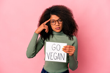 Young african american curly woman holding a go vegan placard pointing temple with finger, thinking, focused on a task.