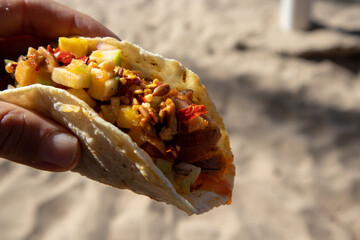 A hand holding a fish taco above a sandy beach in Mexico