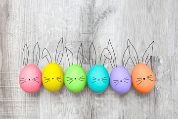 Colorful DIY painted eggs with cute drawing as bunny face and ears on wooden texture background. Creative idea of Easter, a symbol of fun and happy holiday in April. Colored oral row on wood surface.