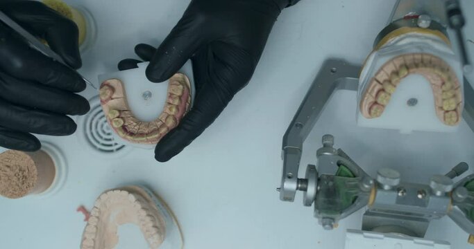 A male technician is wearing black disposable gloves, and cleaning a model of a lower jaw with a metal tool. There is an articulator with a dental prothesis on the table on front of him.