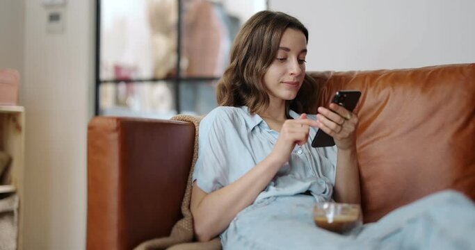 Young and cheerful woman using smart phone at home, sitting on the comfortable sofa in a cozy and stylish living room. Dolly in shot