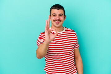 Young caucasian man isolated on blue background winks an eye and holds an okay gesture with hand.