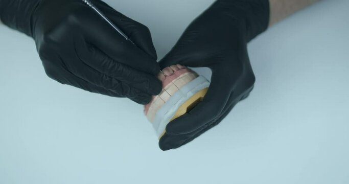 A male dental technician is wearing black disposable gloves and holding a model of a jaw. He is cleaning the space between the teeth of dental prothesis with a sharp metal tool.