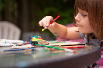 Young Girl paints Rocks outside on Patio Table to be creative outside on a sunny summer day and surrounded by nature