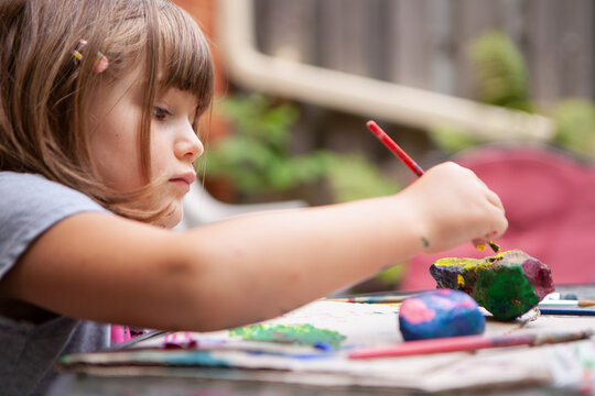 Young Girl paints Rocks outside on Patio Table to be creative outside on a sunny summer day and surrounded by nature