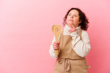 Woman with Down syndrome cooking at home isolated on pink background looking sideways with doubtful and skeptical expression.