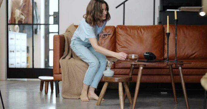 Young woman dressed casually in blue tones feeling comfortable and happy at home, going with coffee, phone, sitting on a leather couch