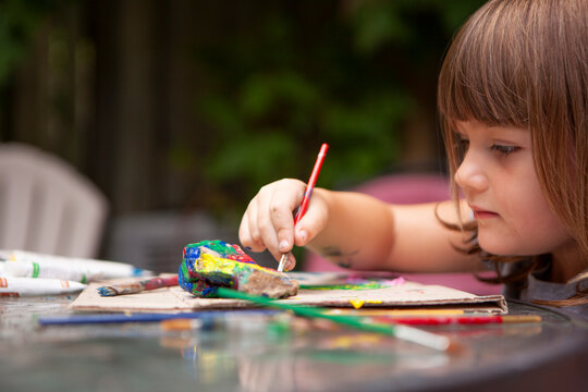 Young Girl paints Rocks with colorful Paints and Brushes outside to be creative outside on a sunny summer day and surrounded by nature