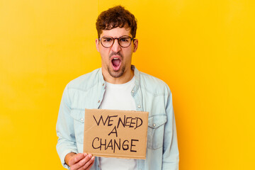 Young caucasian man holding a we need a change placard isolated screaming very angry and aggressive.