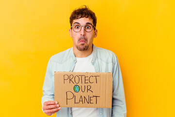 Young caucasian man holding a protect our planet placard isolated shrugs shoulders and open eyes confused.
