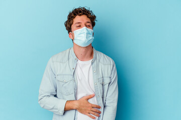 Young caucasian man wearing an antiviral mask isolated on blue background touches tummy, smiles gently, eating and satisfaction concept.