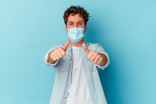 Young Caucasian Man Wearing An Antiviral Mask Isolated On Blue Background With Thumbs Ups, Cheers About Something, Support And Respect Concept.
