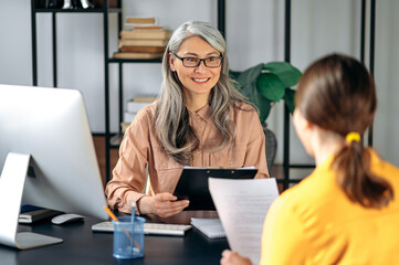 Friendly female mature hr manager listening and looks a resume of a new applicant introduction for a job. Gray-haired female employer talks and meets with female job seeker. - obrazy, fototapety, plakaty