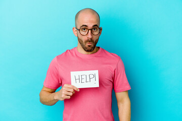 Young caucasian bald man holding a Help placard isolated on purple background shrugs shoulders and open eyes confused.