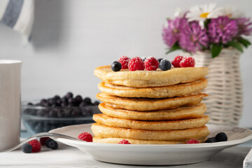 Stack of pancakes with fresh berries on a white plate and a vase of wildflowers on the table