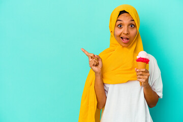 Young muslim woman eating an ice cream isolated on blue background pointing to the side