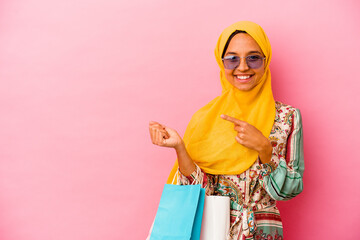 Young muslim woman shopping some clothes isolated on pink background smiling and pointing aside, showing something at blank space.