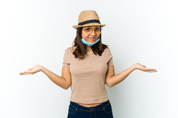 Young latin woman wearing hat and mask to protect from covid isolated on white background doubting and shrugging shoulders in questioning gesture.