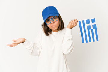 Young hispanic mixed race woman holding a greece flag shrugs shoulders and open eyes confused.