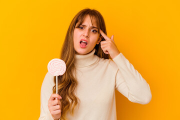 Young caucasian woman holding a lollipop isolated on yellow background showing a disappointment gesture with forefinger.