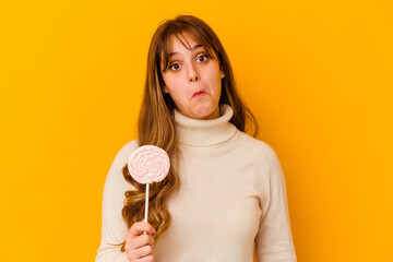 Young caucasian woman holding a lollipop isolated on yellow background shrugs shoulders and open...
