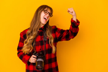 Young photographer caucasian woman isolated on yellow background raising fist after a victory, winner concept.