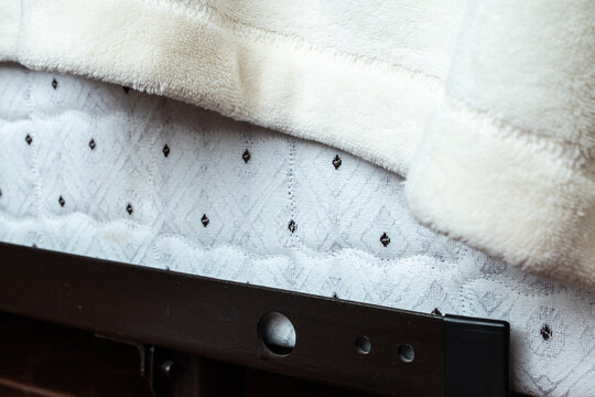 Up close corner of a quilt-patterned bed box spring with black dots on the fabric, covered by a draping fuzzy white blanket on a steel metal frame with adjustable slots, 2021.