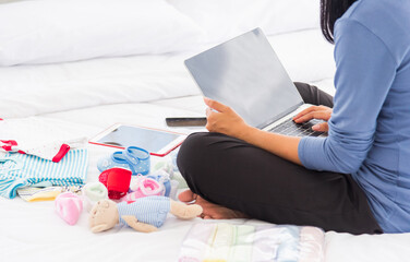 Asian mother shopping online for her baby clothes on laptop computer and tablet on the bed she...