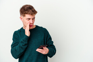 Young caucasian blonde man using a telephone isolated on white background is saying a secret hot...