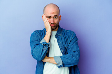 Young caucasian bald man isolated on purple background blows cheeks, has tired expression. Facial expression concept.