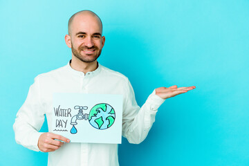 Young caucasian bald man celebrating world water day isolated on blue background showing a copy space on a palm and holding another hand on waist.