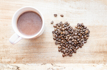 Top view of  Roasted brown coffee beans formed in a heart shape near a cup of hot beverage on wood background, depth of field. The aromatic idea for coffee lovers, a symbol of love on Valentine’s Day.