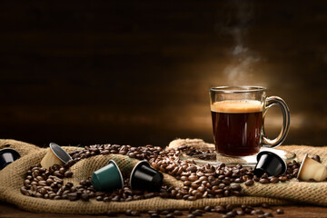 Cup glass of coffee with smoke and coffee beans and coffee capsules on burlap sack on old wooden background