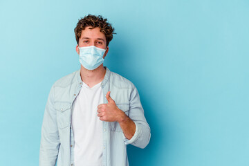 Young caucasian man wearing an antiviral mask isolated on blue background smiling and raising thumb up